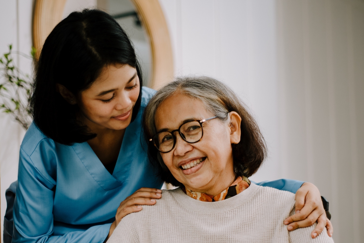 homecare in Lutz An Asian caregiver with short black hair gently wraps her arm around a senior woman’s shoulder, offering comfort. The senior, wearing glasses, looks into the camera with a calm, reassured expression.