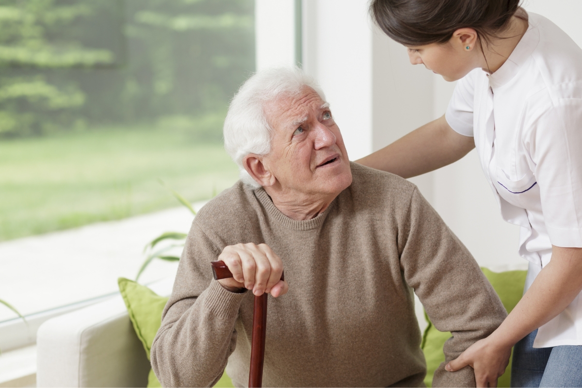 homecare in Putnam Valley An elderly man in a brown sweater looks at an Asian female caregiver helping him rise from a sofa.