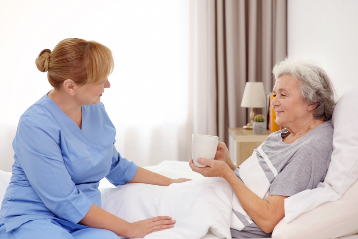 homecare in Riverview (1) A caregiver in blue scrubs with a bun creates a comforting environment for a senior woman lying in bed, holding a cup of tea.