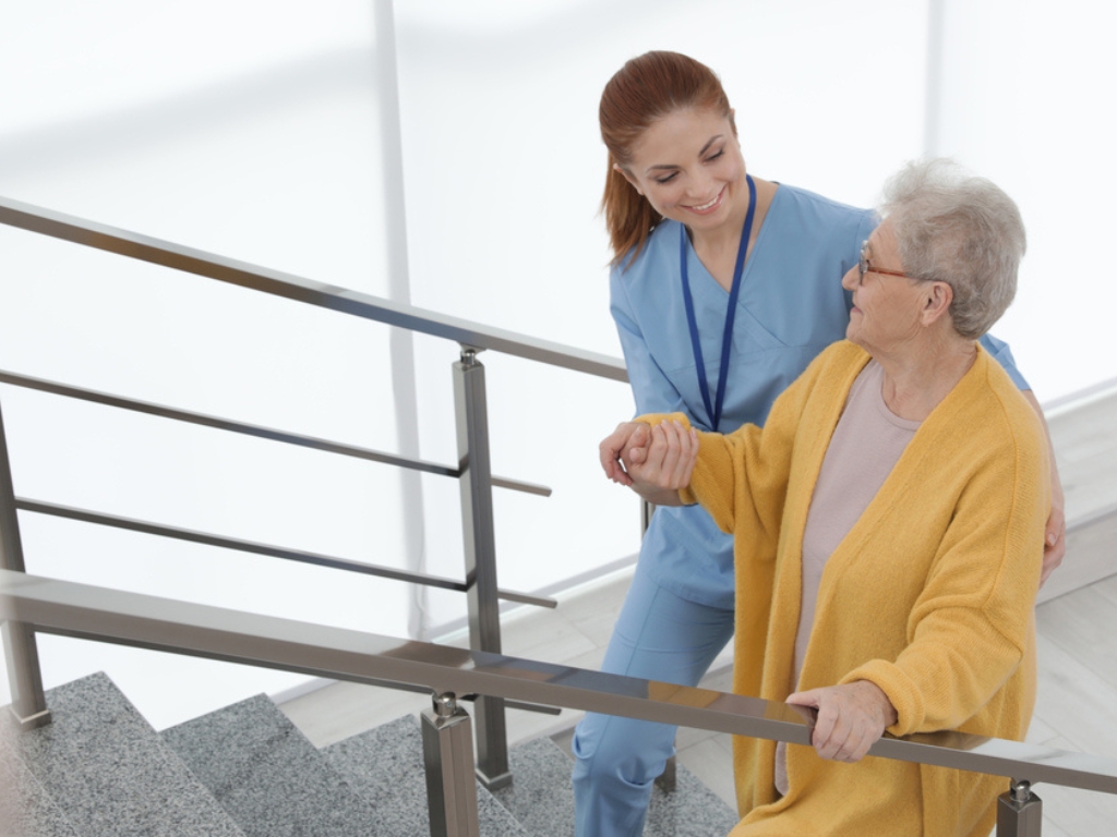 Home Care in Pebble Creek A female caregiver wearing light blue scrubs assists a senior woman in a yellow cardigan as she carefully climbs up the stairs. The caregiver offers steady support, reflecting the attentiveness and safety provided through senior home care in Pebble Creek.