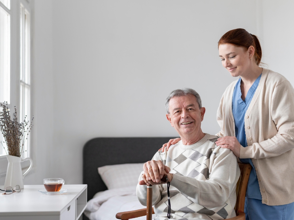 Senior Home Care in Rye, NY A young female caregiver in blue scrubs and a beige cardigan gently looks over a male senior who is seated on a chair, gazing into the camera.