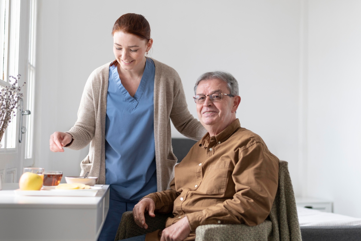 A cheerful female caregiver in scrubs and a cardigan provides assistance to a smiling elderly man seated at a table with tea and fruit, in a bright, cozy room—illustrating compassionate and professional home care in Apollo Beach.