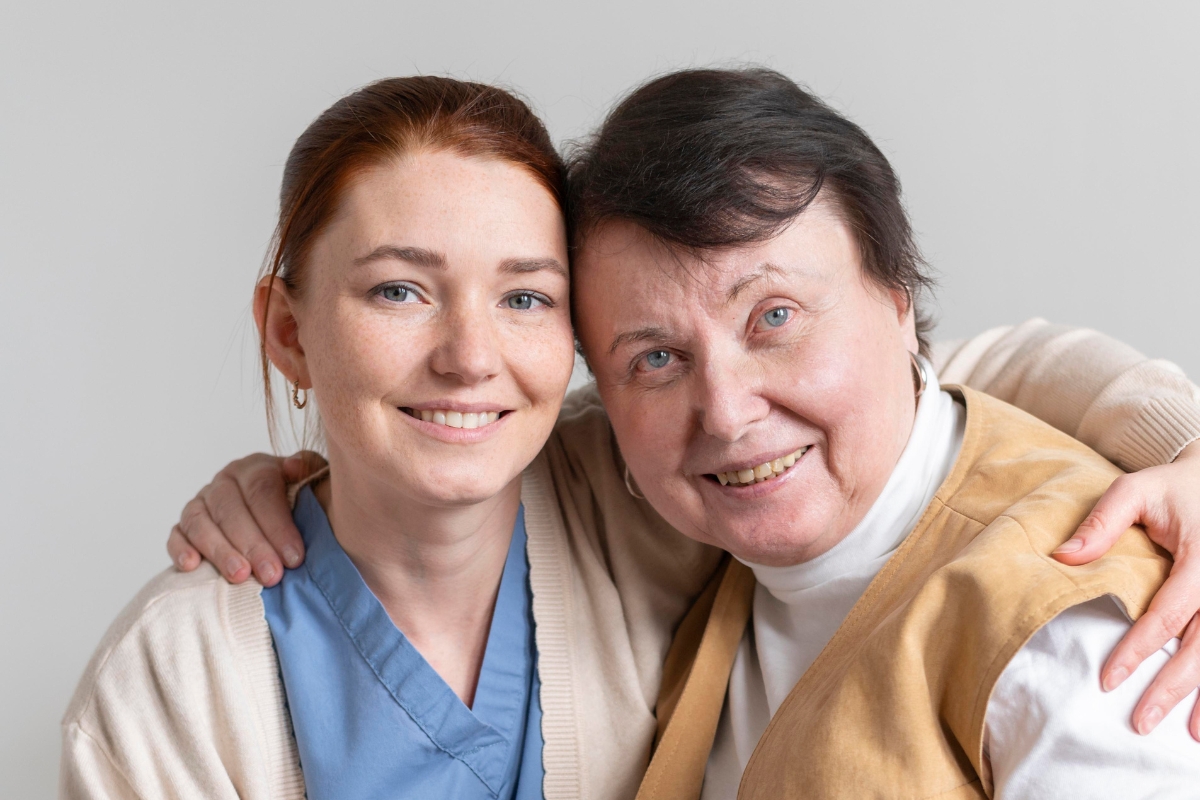 A smiling senior woman and her caregiver share a warm embrace, symbolizing trust and companionship. The caregiver is dressed in scrubs and a cardigan, while the senior wears a tan vest and white turtleneck. This heartwarming moment reflects the compassionate and personalized senior home care in Larchmont offered by dedicated professionals.
