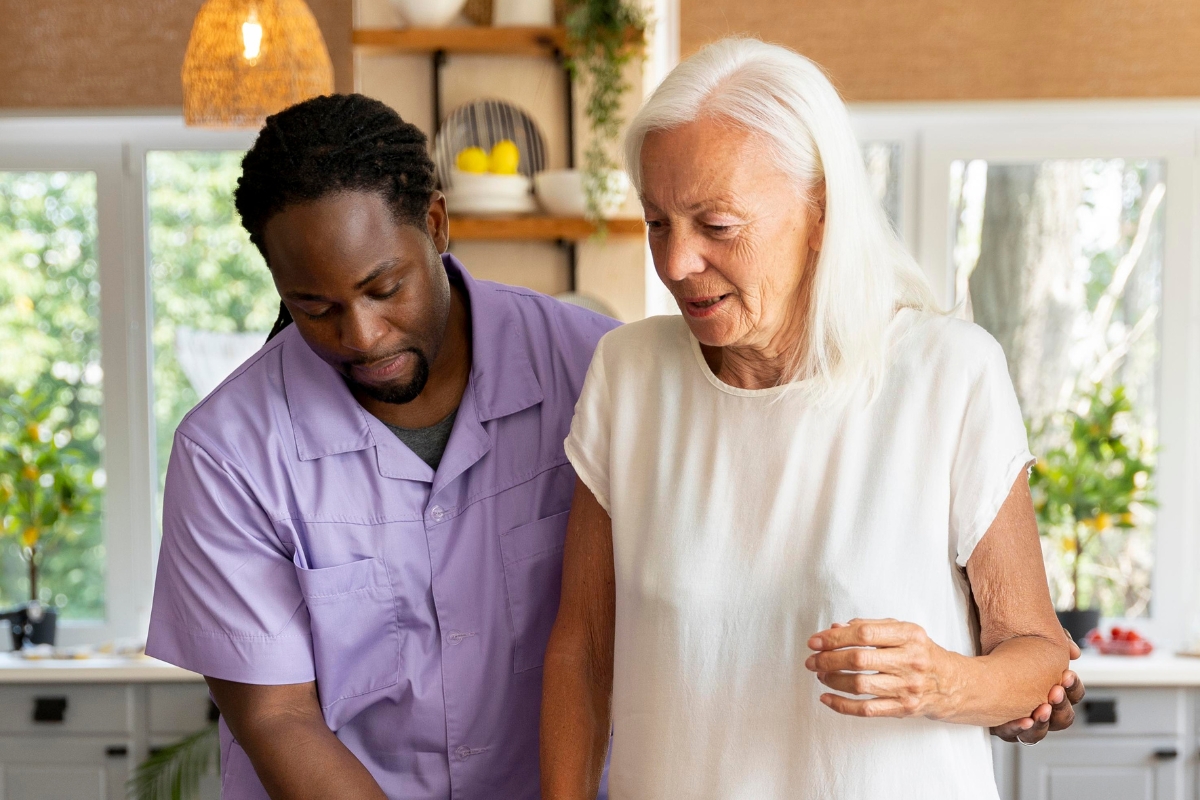 A caregiver in a lavender uniform assists an elderly woman with white hair as they walk through a bright, modern kitchen. This image represents compassionate senior care in White Plains.
