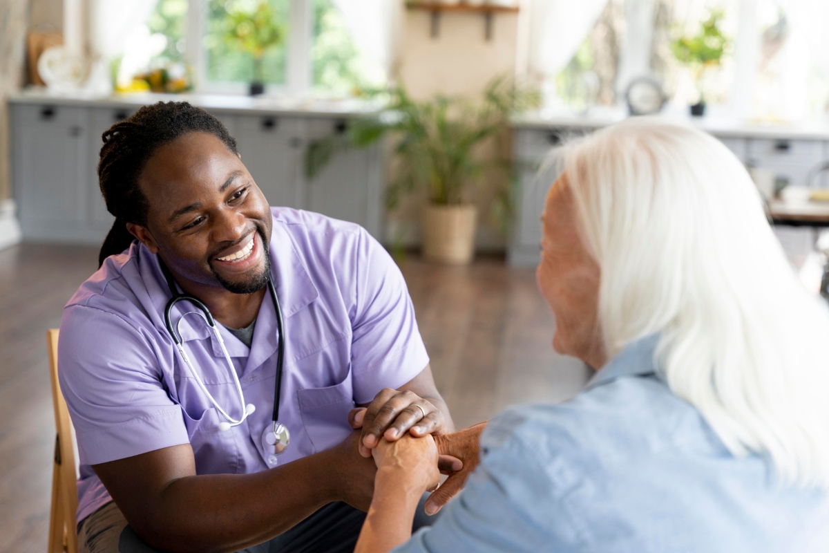 : A cheerful male caregiver in a purple uniform with a stethoscope smiles warmly while holding hands with a senior woman in a cozy, well-lit home setting. This image reflects compassionate and supportive home care in Cheval.