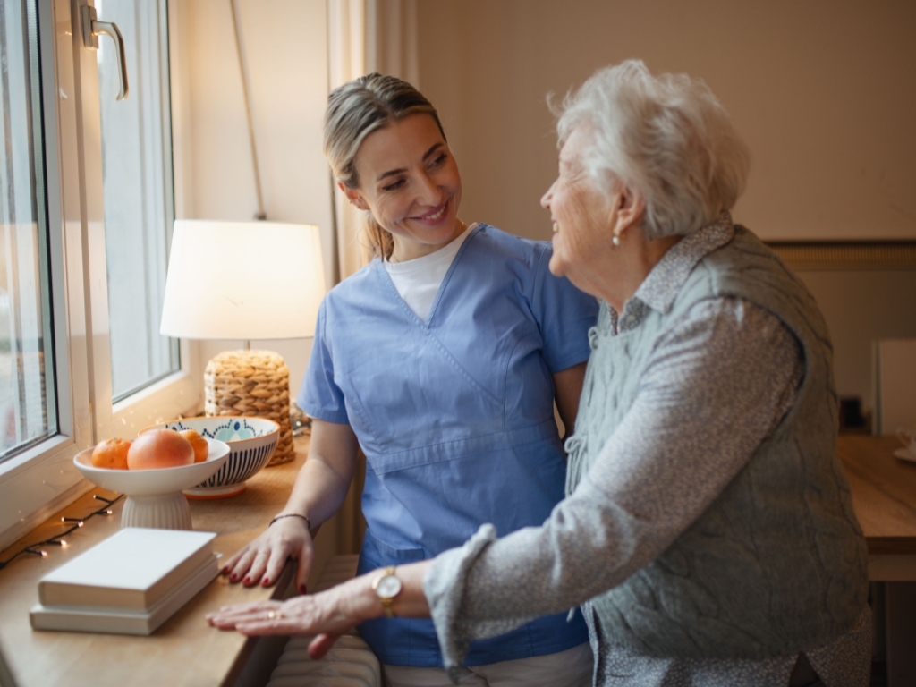 : A smiling caregiver in blue scrubs provides compassionate support to an elderly woman near a bright window, symbolizing trusted home care in Connecticut.