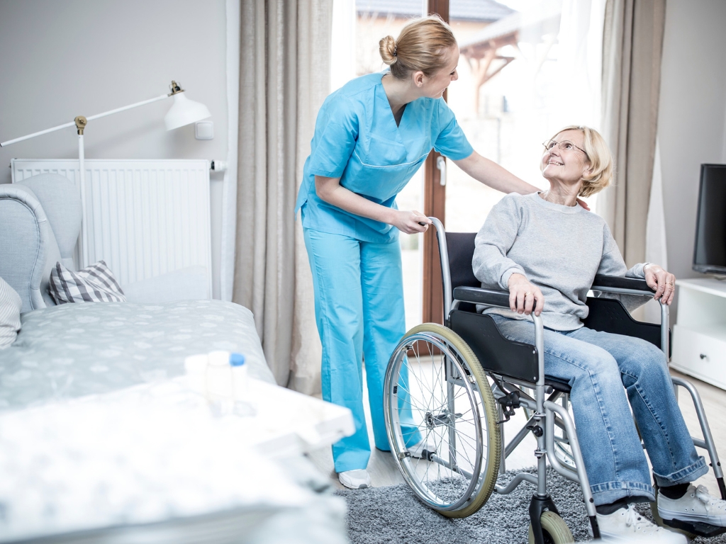A caregiver in blue scrubs assists a smiling senior woman in a wheelchair inside a bright, comfortable home setting, symbolizing compassionate home care in North Dade.