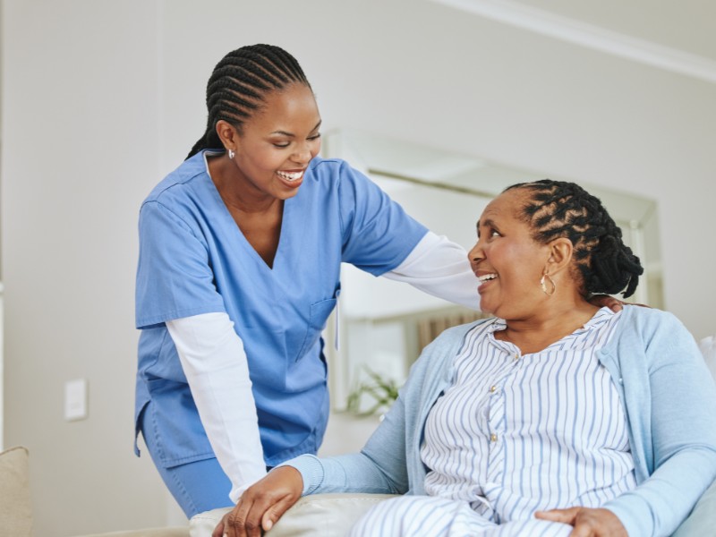 A cheerful caregiver resting a gentle hand on the shoulder of a smiling older woman sitting on a couch, representing trustworthy Respite Care in Temple Terrace.