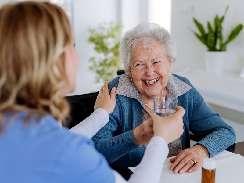 A caring professional sharing a warm laugh and handing a glass of water to an older woman, providing gentle Medication Supervision Temple Terrace.